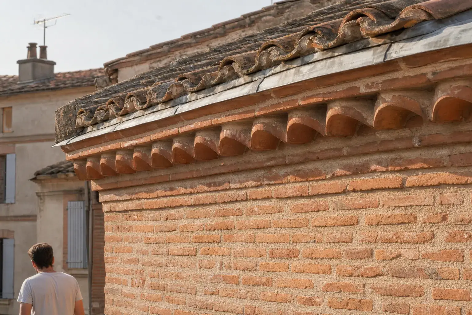 Rive d’une maison toulousaine : mur de brique foraine rose, génoise de deux rangs de tuiles canal en encorbellement, versant patiné au-dessus